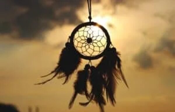 Shadowed dreamcatcher with feather tassels, a beautiful nod to traditions & customs, hangs against a sunset sky.