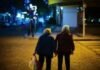 Two elderly people walk hand in hand on a dimly lit street at night, with one carrying a plastic bag, savoring these tender moments as though they were your last days.