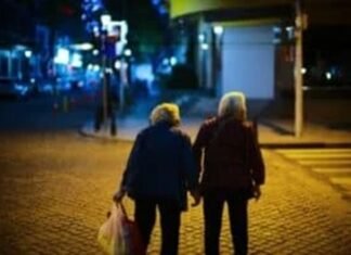 Two elderly people walk hand in hand on a dimly lit street at night, with one carrying a plastic bag, savoring these tender moments as though they were your last days.