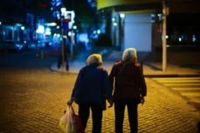 Two elderly people walk hand in hand on a dimly lit street at night, with one carrying a plastic bag, savoring these tender moments as though they were your last days.