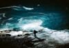 A person stands inside a large ice cave with illuminated blue walls and a rocky ground, feeling an odd sense of calm despite the potentially bad situation.