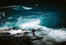 A person stands inside a large ice cave with illuminated blue walls and a rocky ground, feeling an odd sense of calm despite the potentially bad situation.