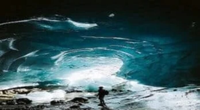 A person stands inside a large ice cave with illuminated blue walls and a rocky ground, feeling an odd sense of calm despite the potentially bad situation.