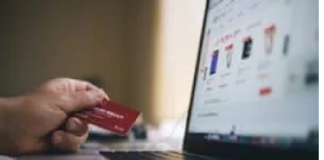 A person holds a red credit card, determined to get out of debt, while shopping online on a laptop displaying product listings.