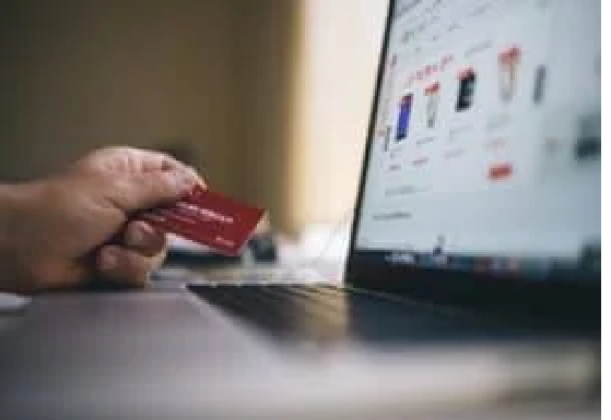 A person holds a red credit card, determined to get out of debt, while shopping online on a laptop displaying product listings.