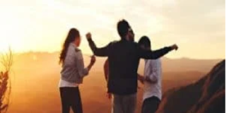 Four people stand on a hilltop at sunset, arms raised, overlooking a scenic view. How does your life shape your age