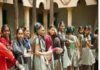 A group of middle class school students in uniform stands in a line outdoors, holding items in their hands, with trees and a building in the background.