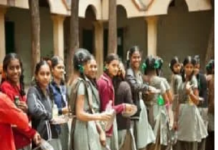 A group of middle class school students in uniform stands in a line outdoors, holding items in their hands, with trees and a building in the background.