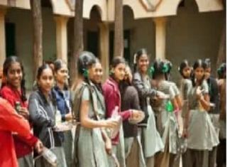 A group of middle class school students in uniform stands in a line outdoors, holding items in their hands, with trees and a building in the background.