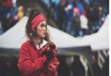 A person wearing a red sweatshirt and headband stands outdoors, holding red pom-poms. The background features tents and an indistinct crowd. Stay healthy.