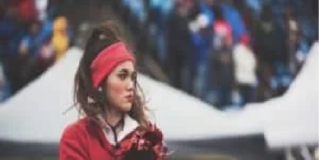 A person wearing a red sweatshirt and headband stands outdoors, holding red pom-poms. The background features tents and an indistinct crowd. Stay healthy.