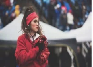 A person wearing a red sweatshirt and headband stands outdoors, holding red pom-poms. The background features tents and an indistinct crowd. Stay healthy.