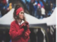 A person wearing a red sweatshirt and headband stands outdoors, holding red pom-poms. The background features tents and an indistinct crowd. Stay healthy.