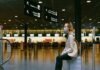 A person wearing a mask sits on a suitcase in an airport terminal, surrounded by directional signs, as reminders of the Corona Covid-19 era linger near the check-in counters.