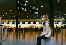 A person wearing a mask sits on a suitcase in an airport terminal, surrounded by directional signs, as reminders of the Corona Covid-19 era linger near the check-in counters.