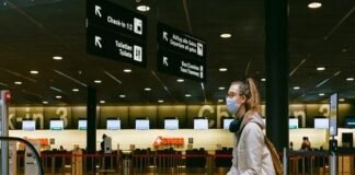 A person wearing a mask sits on a suitcase in an airport terminal, surrounded by directional signs, as reminders of the Corona Covid-19 era linger near the check-in counters.