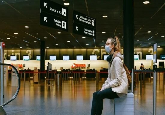 A person wearing a mask sits on a suitcase in an airport terminal, surrounded by directional signs, as reminders of the Corona Covid-19 era linger near the check-in counters.