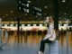 A person wearing a mask sits on a suitcase in an airport terminal, surrounded by directional signs, as reminders of the Corona Covid-19 era linger near the check-in counters.