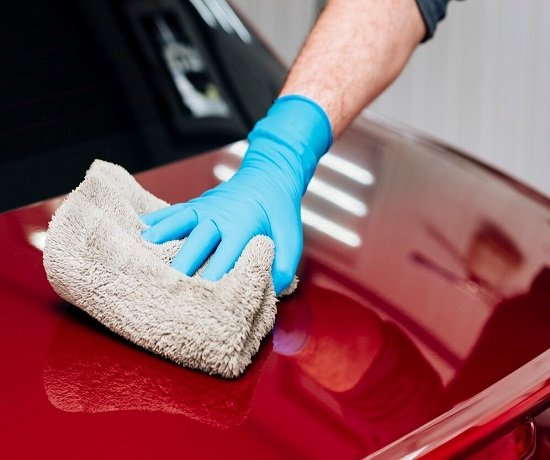A person wearing blue gloves is meticulously wiping a red car's surface with a grey cloth—a simple yet effective touch that could be the cornerstone of your next Car Wash Franchise Idea.