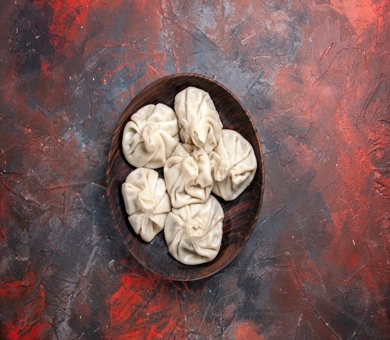 A wooden bowl filled with steamed dumplings is placed on a textured, red and gray surface.