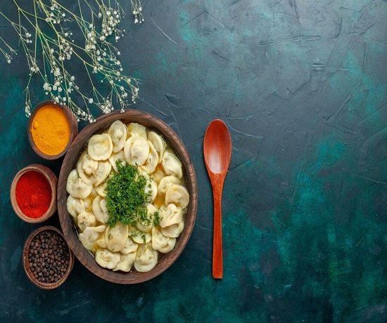 A bowl of ravioli garnished with herbs and accompanied by three small bowls of spices and a wooden spoon on a dark green textured surface.