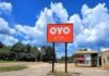 A red sign with "OYO Hotel" stands in front of a parking lot, with trees and a blue sky with clouds in the background.