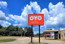 A red sign with "OYO Hotel" stands in front of a parking lot, with trees and a blue sky with clouds in the background.