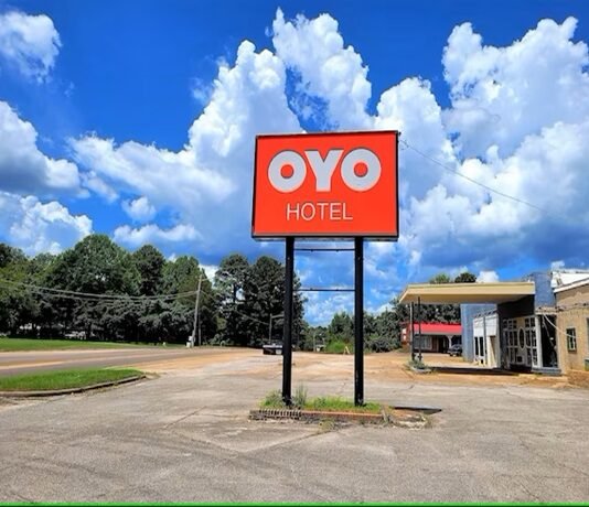 A red sign with "OYO Hotel" stands in front of a parking lot, with trees and a blue sky with clouds in the background.