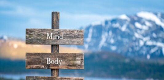 A wooden sign with "Mind," "Body," "Connection" stands gracefully against a serene lake and mountain backdrop, capturing the essence of natural health.