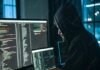 A person in a hoodie works on multiple computer screens displaying code in a dimly lit room, dark web hinting at illegal activity.