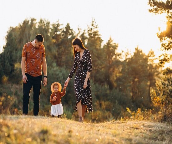 A couple and their child walk hand in hand on a grassy field, embodying the essence of family relationships. Trees stand tall in the background as the sun sets, casting a warm glow that radiates well-being over the serene scene.