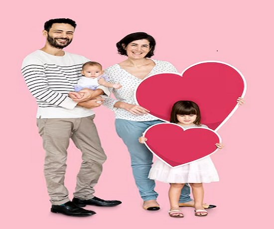 A family of four, radiating well-being, stands together against a pink background. The father cradles a baby while the mother stands beside a young girl holding two large pink heart shapes, showcasing their strong family relationship.