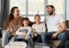 A smiling family of four sits on a couch, laughing together, embodying the essence of a strong family relationship. Two children are seated in the middle, enveloped by the warmth and well-being provided by their parents on either side.