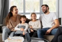 A smiling family of four sits on a couch, laughing together, embodying the essence of a strong family relationship. Two children are seated in the middle, enveloped by the warmth and well-being provided by their parents on either side.