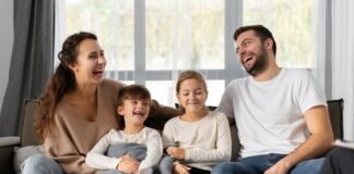 A smiling family of four sits on a couch, laughing together, embodying the essence of a strong family relationship. Two children are seated in the middle, enveloped by the warmth and well-being provided by their parents on either side.