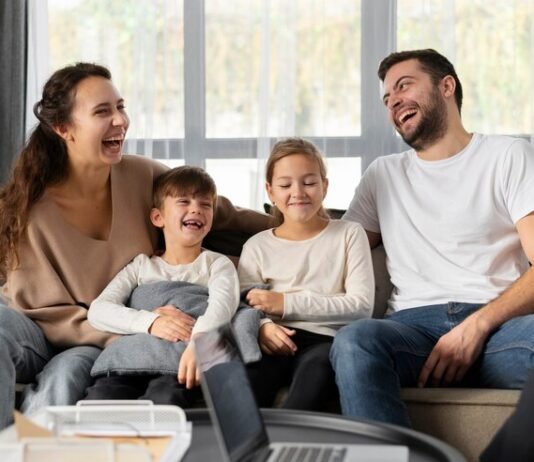 A smiling family of four sits on a couch, laughing together, embodying the essence of a strong family relationship. Two children are seated in the middle, enveloped by the warmth and well-being provided by their parents on either side.