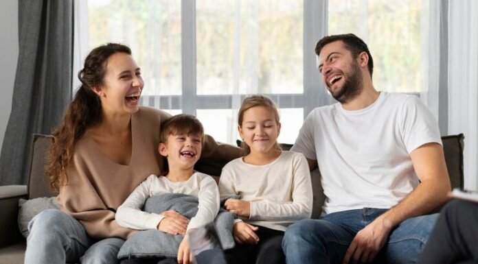 A smiling family of four sits on a couch, laughing together, embodying the essence of a strong family relationship. Two children are seated in the middle, enveloped by the warmth and well-being provided by their parents on either side.