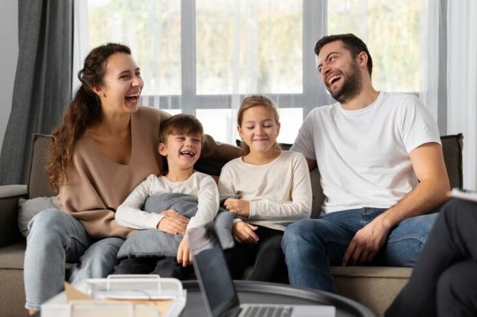 A smiling family of four sits on a couch, laughing together, embodying the essence of a strong family relationship. Two children are seated in the middle, enveloped by the warmth and well-being provided by their parents on either side.