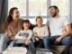 A smiling family of four sits on a couch, laughing together, embodying the essence of a strong family relationship. Two children are seated in the middle, enveloped by the warmth and well-being provided by their parents on either side.