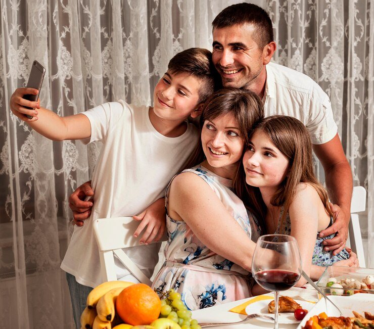 A family of four, cherishing their well-being and strong family relationship, takes a selfie at a dining table adorned with a vibrant variety of fruit and a glass of red wine in the foreground.