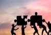 Silhouettes of people working together to fit giant puzzle pieces against a sunset background, embodying the spirit of challenges and personality development.