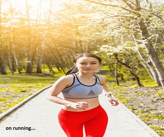 A woman in red leggings and a gray sports bra jogs along a sunny path through a park, surrounded by greenery and trees, focusing on her well-being.