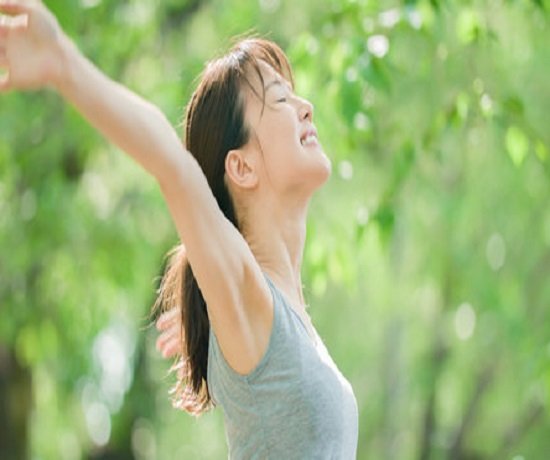 A person outdoors with eyes closed, arms outstretched, reveling in the well-being that comes from fresh air. Trees and greenery flourish in the background.