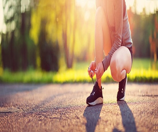 Person tying running shoe laces on a paved path outdoors, with sunlight filtering through trees in the background, embracing a moment of well-being.