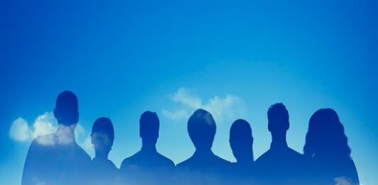 Silhouettes of seven people, representing teamwork, overlay a city skyline under a blue sky with clouds.