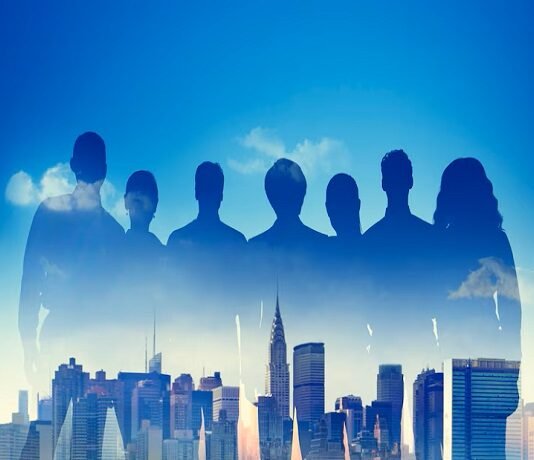 Silhouettes of seven people, representing teamwork, overlay a city skyline under a blue sky with clouds.