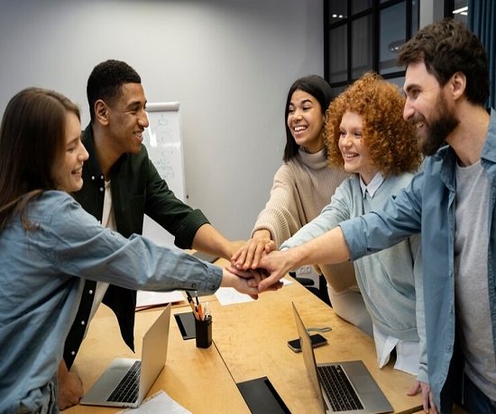 A diverse group of five people in casual attire stands around a table with laptops, smiling as they stack their hands together, showcasing the essence of teamwork in an office setting.