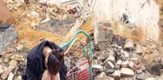 A person sits hunched over amidst rubble and debris, remnants of an earthquake bomb or airstrike, with hoses and broken bricks scattered in the background.