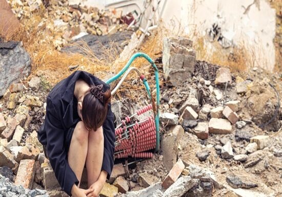 A person sits hunched over amidst rubble and debris, remnants of an earthquake bomb or airstrike, with hoses and broken bricks scattered in the background.