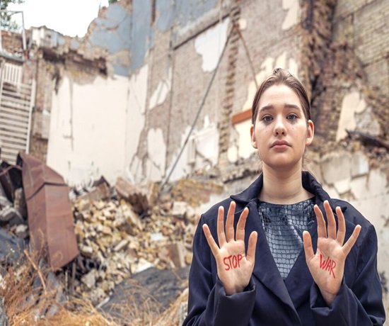 In front of a pile of rubble from a destroyed building, a person stands with "Stop War" written on their hands, symbolically opposing the devastation left in the wake of an airstrike.